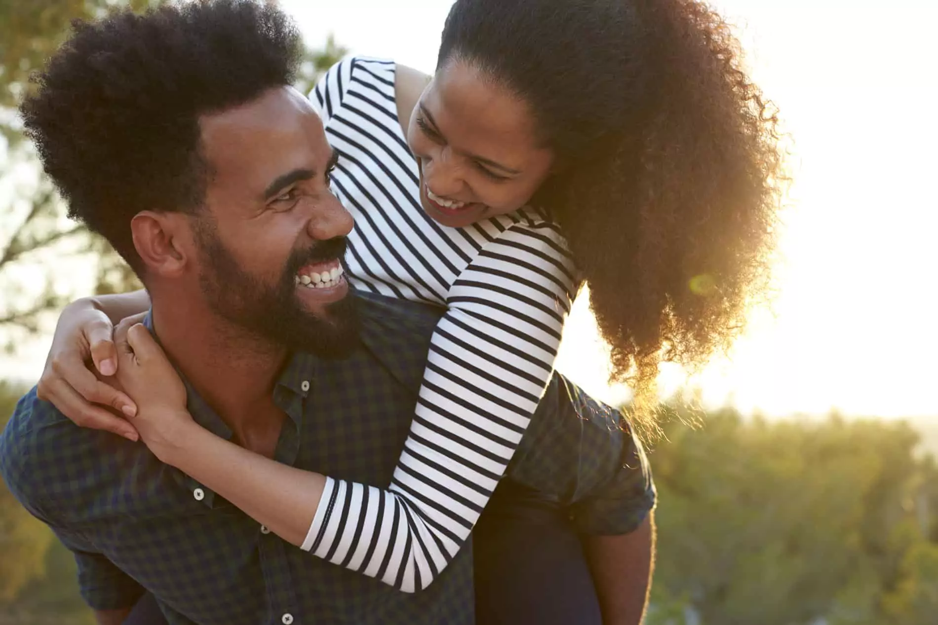 A couple shares a playful moment, embodying how to bring humor into your marriage. The man smiles widely as the woman playfully leans on his back, both laughing together in a lighthearted and affectionate manner.