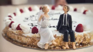 Photo of bride and groom cake toppers sitting barefoot at the edge of their cake, resting after planning their wedding.