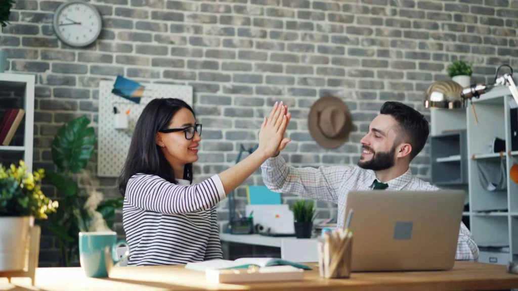 A couple sits at a desk high-fiving each other, representing teamwork and success, illustrating how to get on the same financial page as your spouse.