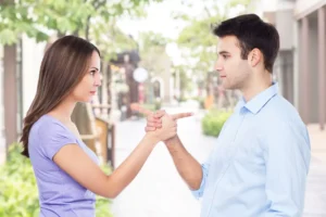 Two people, a man and a woman, are standing outdoors, facing each other with serious expressions, pointing fingers at each other as if they are engaging in a blame game.