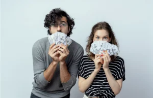 A couple holding stacks of cash in front of their faces, symbolizing a lighthearted yet important conversation about how to talk about budgeting in a relationship.