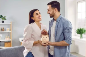 A smiling couple holds a piggy bank together, symbolizing their shared approach to managing money and finances in their marriage.