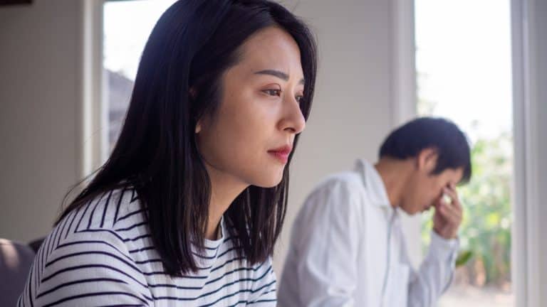 Troubled couple sitting on couch. She's in the foreground with a blank stare, he's in the back, wiping tears with his hand.