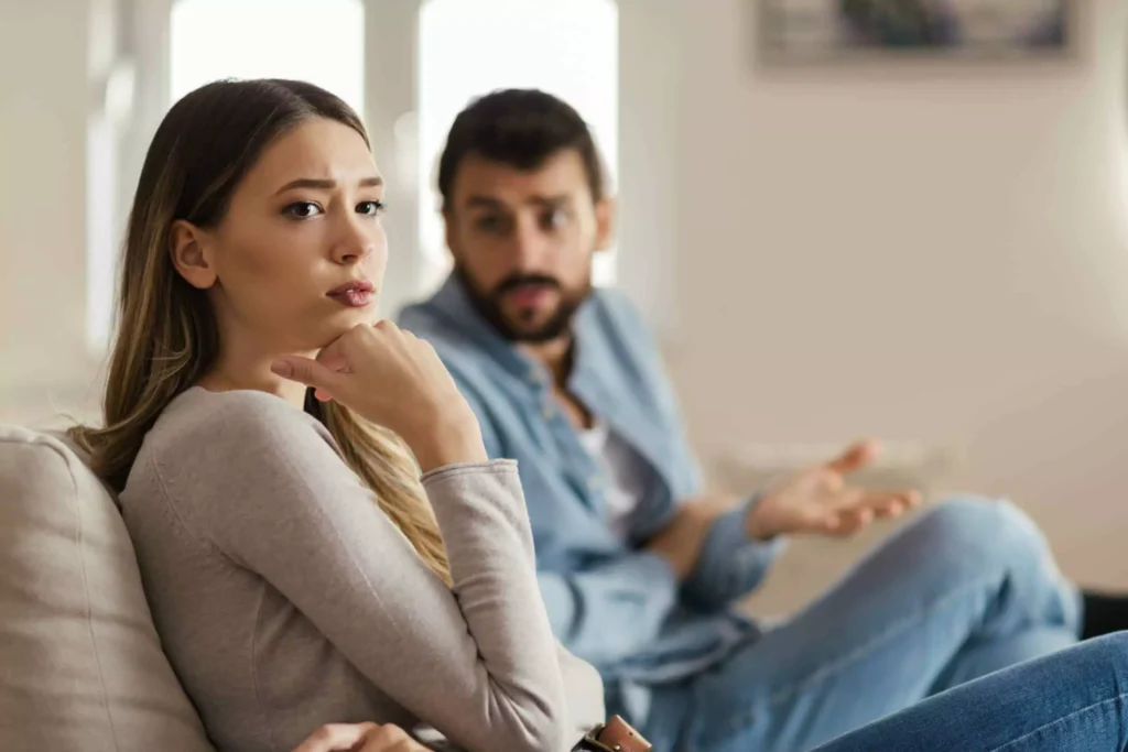 A woman sitting on a couch with a thoughtful expression, looking away from a man in the background who appears to be talking. This scene could represent red flags in a relationship, where communication might be strained or there could be underlying issues.