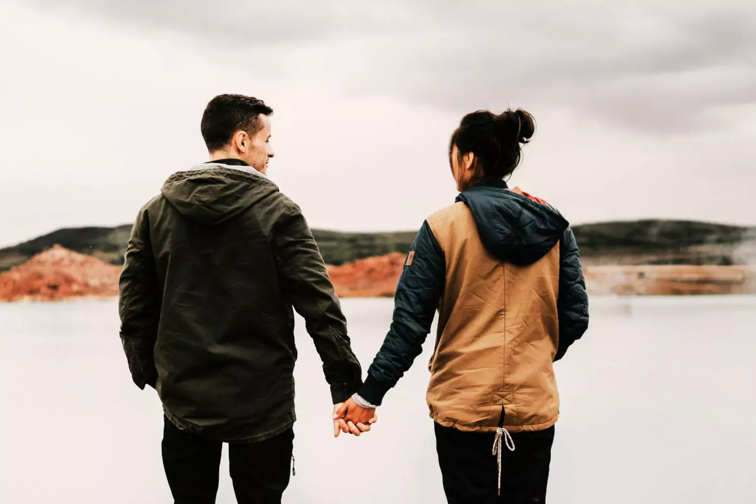 A couple stands hand in hand by a quiet lake under a cloudy sky, reflecting on whether to end a relationship