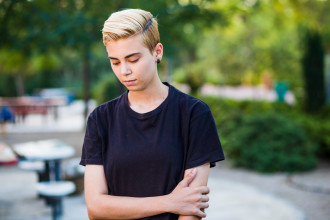 A gender-ambiguous teen standing outside, looking down in dismay
