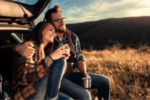 A couple enjoying a cozy date night outdoors, sitting together in the trunk of their car, smiling and holding metal cups as the sun sets over a peaceful, golden landscape.