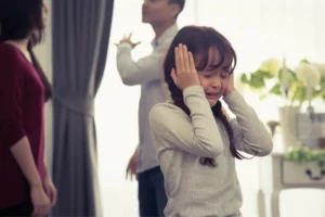 Mother and father fighting in the background. Their little girl covering her ears and crying.