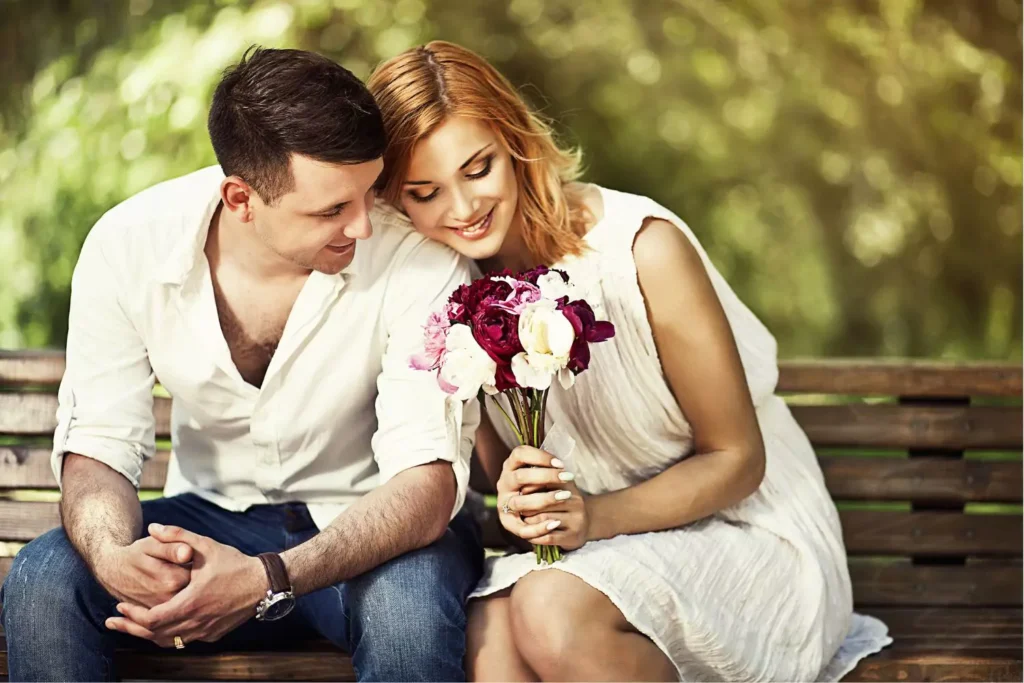 A husband and wife sit on a park bench. The wife is holding a beautiful bouquet of flowers and leaning into her husband's shoulder, depicting authentic intimacy in their marriage.