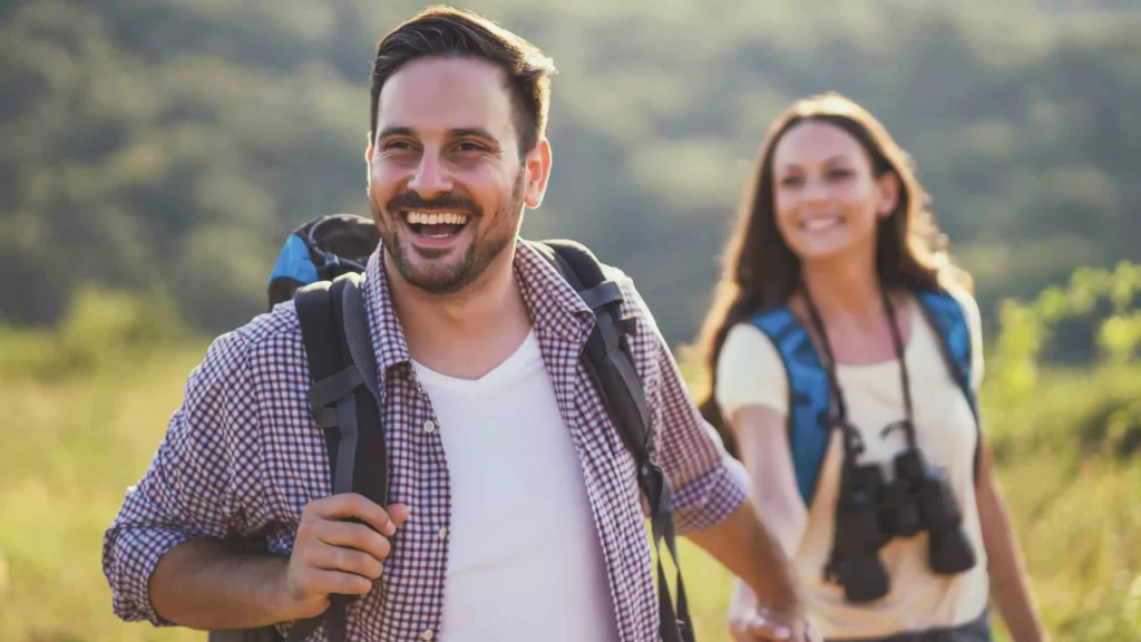 A happy couple is seen hiking together in a scenic, nature-filled setting, wearing backpacks and smiling brightly. The man in the foreground laughs joyfully, while the woman follows closely behind, looking at him with a smile. They appear to be enjoying a shared adventure, a symbol of partnership, exploration, and fun—highlighting ways to have a great marriage, such as spending quality time outdoors, enjoying shared activities, and maintaining a positive attitude together. This image conveys a sense of togetherness, communication, and appreciation for each other, essential components of a thriving relationship.