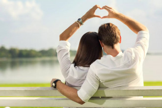A couple sitting on a bench by the water, forming a heart shape with their hands, symbolizing the impact of marriage help in rekindling love and connection.