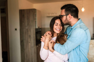 Husband embracing his wife from the side. He's kissing her head, she's smiling and enjoying it.