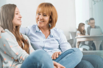 Mom and teen sitting on couch and chatting while dad and younger brother work at the table in the background