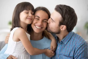 A woman smiles as she is hugged by her daughter and kissed by her husband, capturing the delicate balance of being both a wife and a mom.