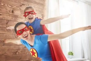 A mother and daughter dressed as superheroes are playing together, embodying bold and courageous parenting. The mother carries the daughter on her back, both with arms outstretched as if flying.