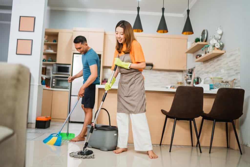 A young couple sweep the floor together in their kitchen. When you share the chores in your marriage and your goal is to out-serve each other, you can bring an end to the chore wars in your home.