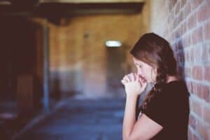 A woman leans against a wall with her eyes closed and hands folded