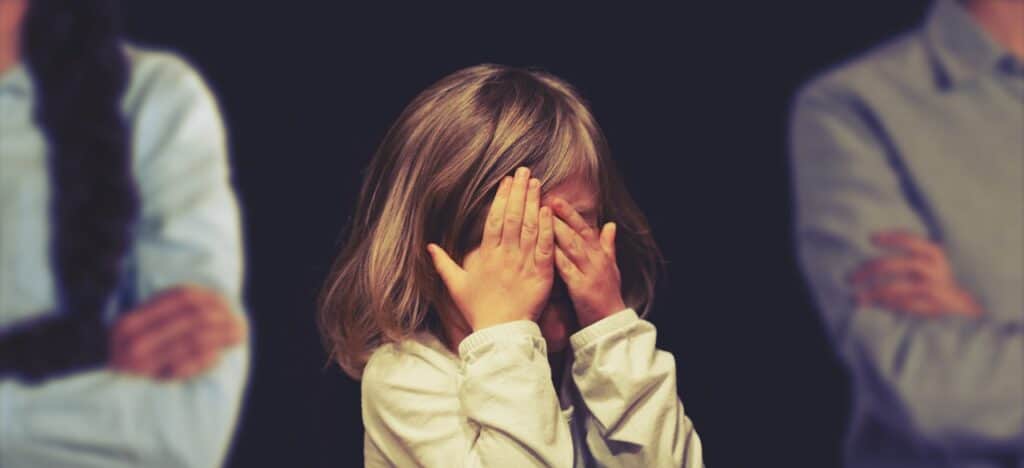 child covering face and parents' stand in background with arms folded