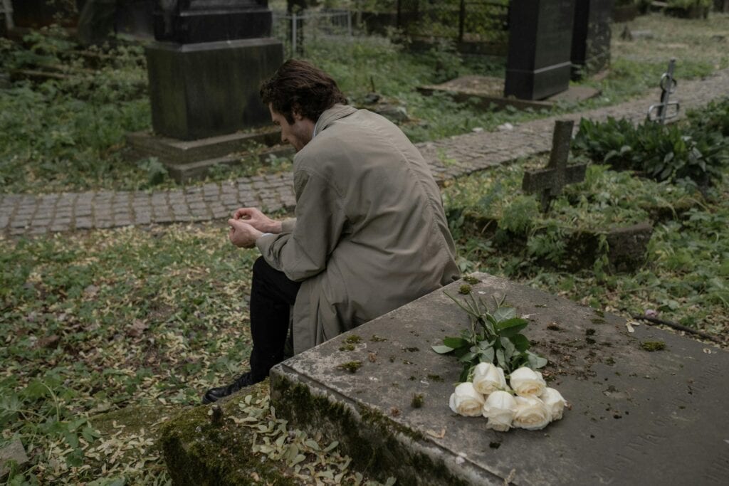 man with back turned sitting by gravestone with flowers