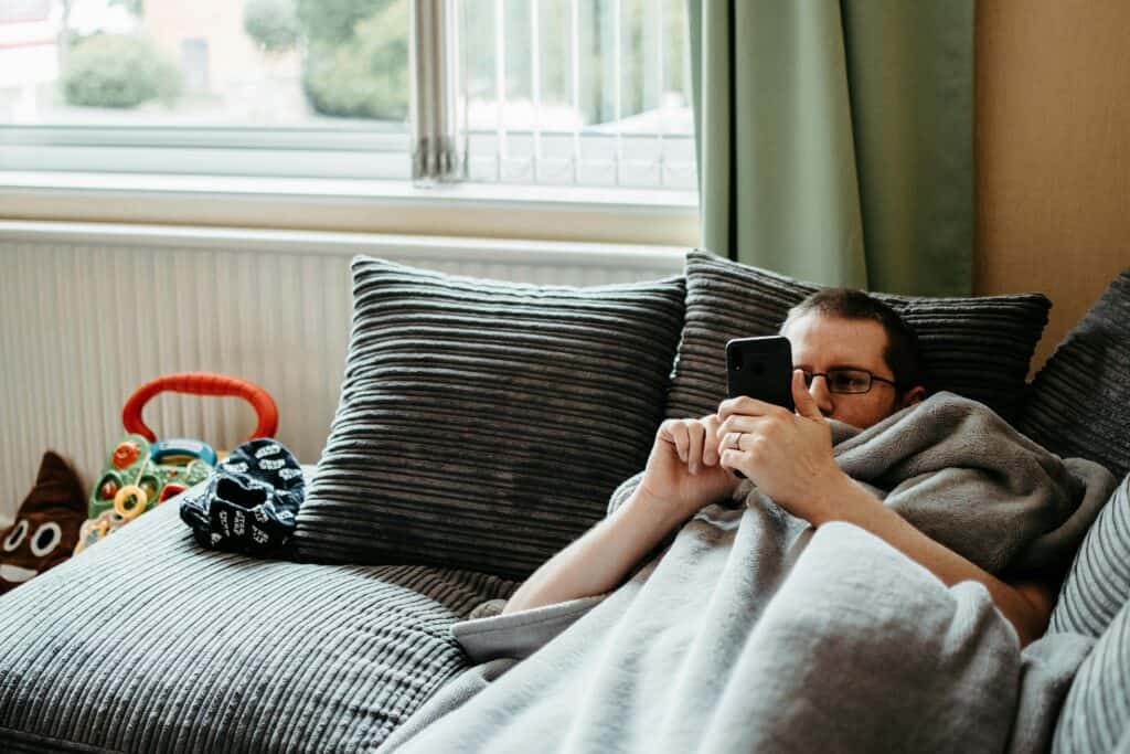 man lying on couch under blanket looking at phone