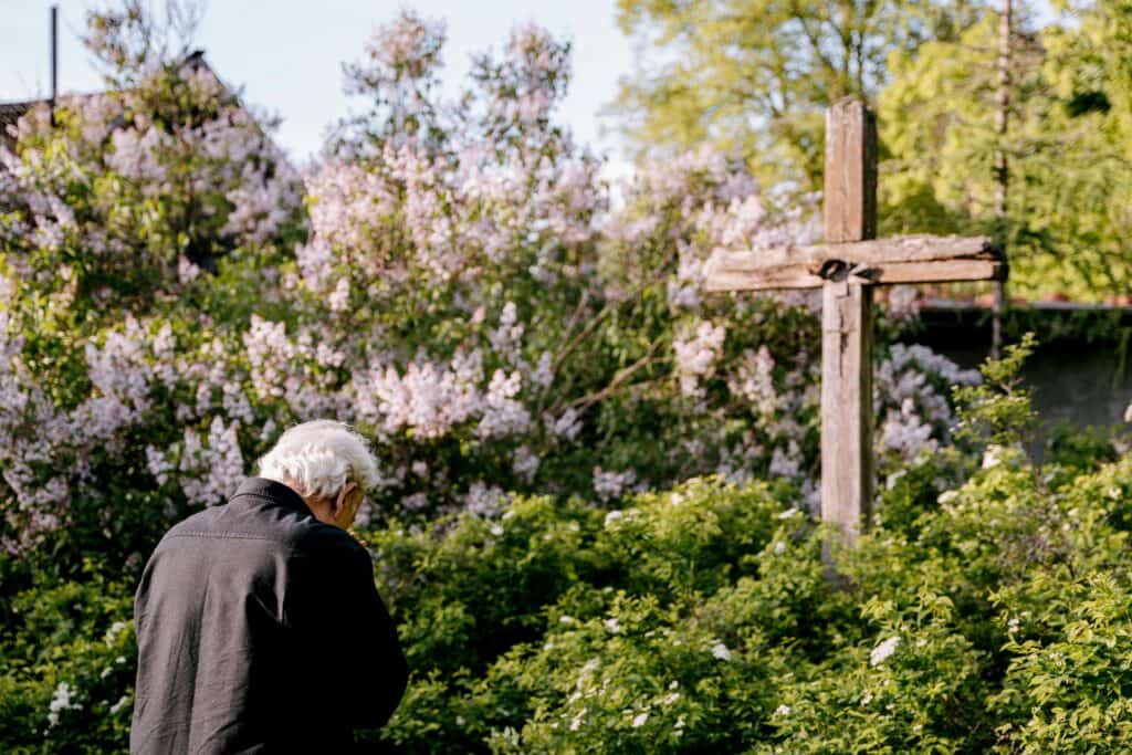 elderly man bowed head near flower trees wooden cross