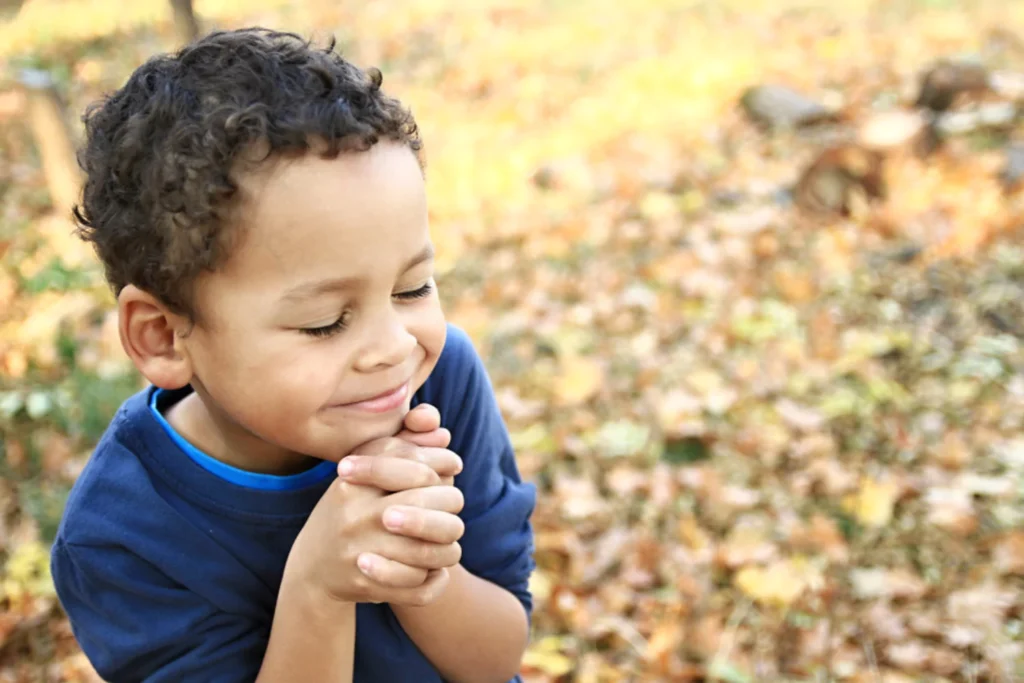 A young boy with curly hair and a blue shirt is praying with his eyes closed and hands clasped, surrounded by autumn leaves. This image captures a moment of teaching kids to pray.