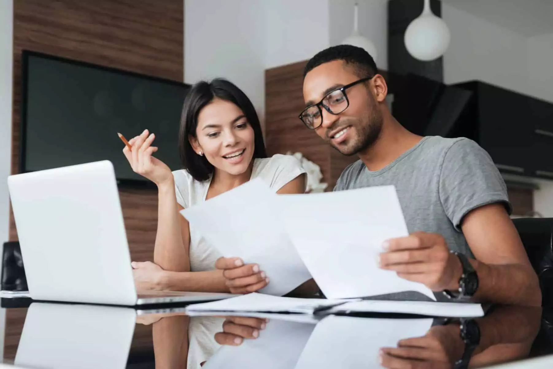 A couple smiles while reviewing documents together at a table with a laptop, highlighting the importance of managing money as a couple