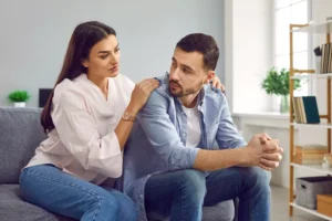 A photo of a husband and wife sitting on a couch, with the wife comforting her husband, who is obviously stressed. If you're the wife of a stressed-out husband, maybe these tips will help you support your husband through work stress.