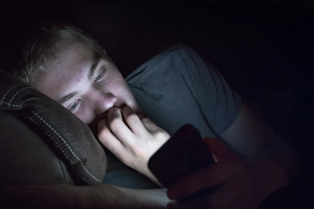 teenaged boy laying in bed looking at porn on his phone in the dark