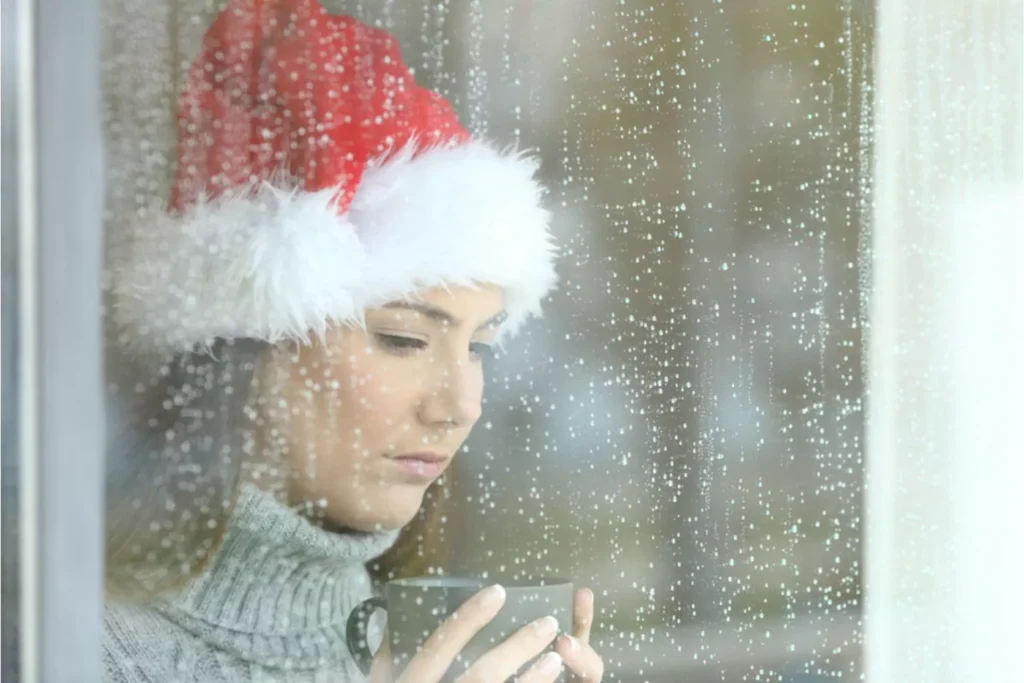 Post-holiday depression can show up as sadness felt by this woman in a holiday hat as she stares out the window at the rain.