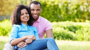 A couple sits in a green field with trees in the background, with the husband's arms wrapped around the wife. They are smiling, as they have been enriching their marriage with devotionals for married couples.