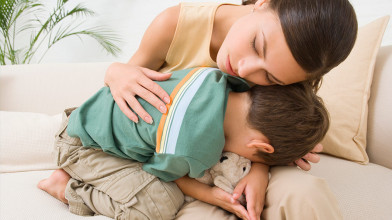 A mom sitting on a couch, either comforting or cuddling with her young son who’s got his face buried in her lap