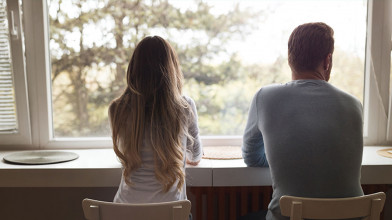 Shown from behind, a seated couple looking out their home window