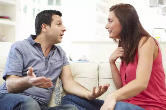 A husband and wife sit on a couch having a serious conversation, reflecting the strain caused by a husband’s selfishness.