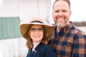Bart Millard and his wife smile warmly in an outdoor setting, dressed in stylish yet cozy attire.