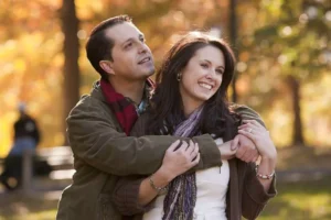 A husband stands behind his wife with his arms around her, both smiling, with fall leaves in the background, conveying the importance of making your spouse feel cherished.