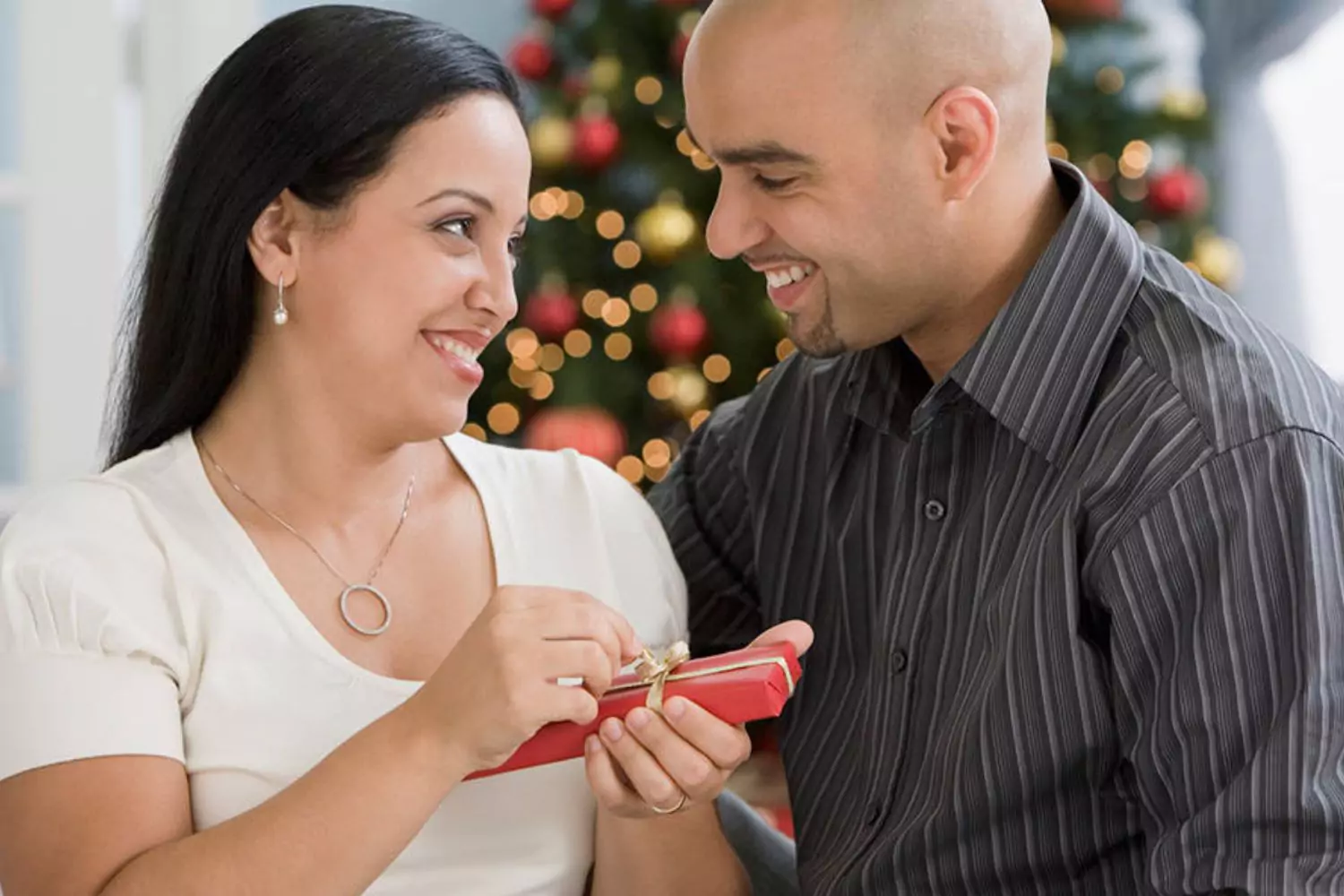 A couple exchanges a Christmas gift in a cozy setting. The woman, smiling warmly, opens a small, neatly wrapped gift from the man. Behind them is a decorated Christmas tree with festive lights. They have learned ways to avoid the stress of Christmas gift giving.