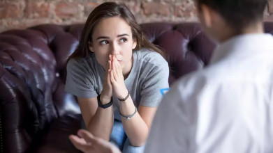 A young woman sitting on a couch, appearing deep in thought and concern, representing the concept of defining trauma.