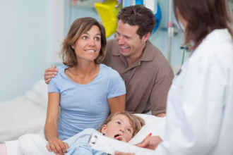 Happy parents receiving good news from a doctor about their daughter who’s lying next to them in a hospital bed after a health scare.