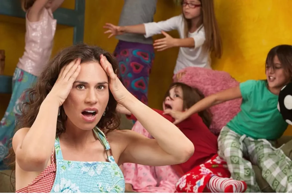 A stressed mother holding her head as children play chaotically in the background, symbolizing challenges of learning how to stop yelling at your kids.