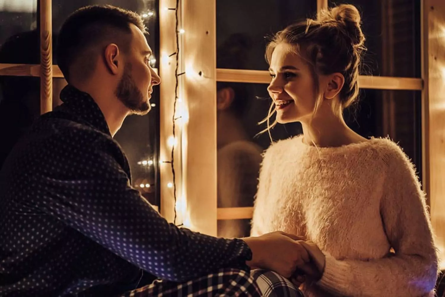 A couple sits by a window adorned with holiday lights, holding hands and smiling warmly, symbolizing the importance of managing expectations in marriage during the holiday season.