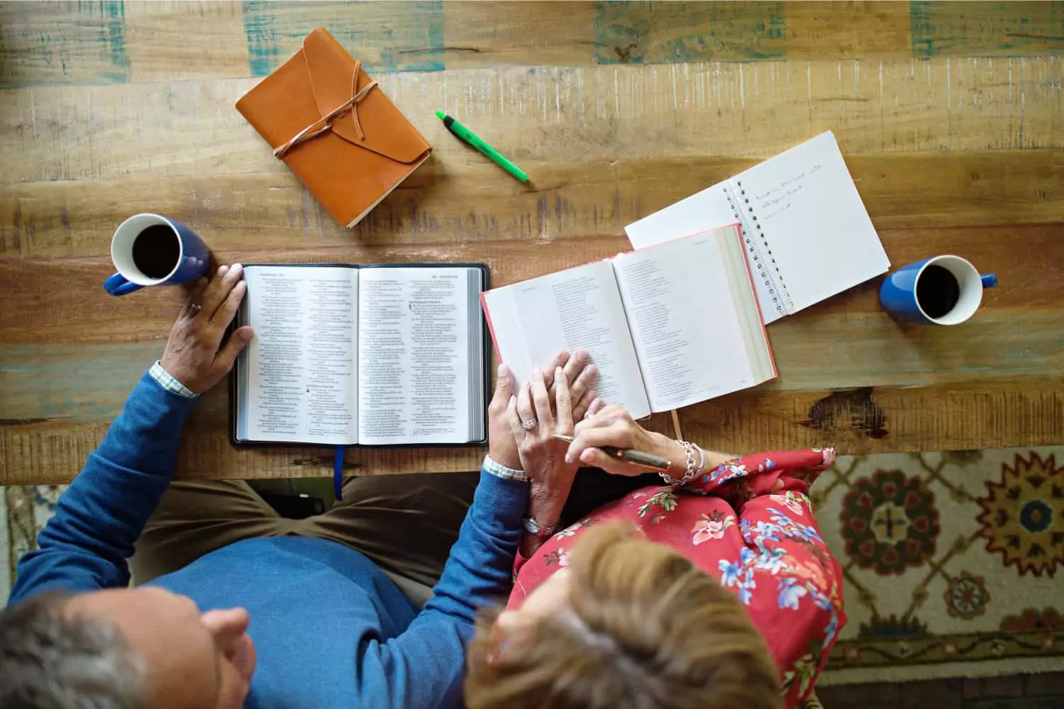 A couple sits together at a rustic wooden table, holding hands while engaging in Bible reading with their spouse, with open books, coffee mugs, and a journal nearby.