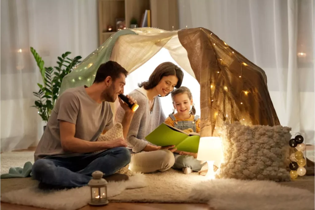 Parents motivate their child to read by reading with their child in an indoor reading tent. Mom and dad reading together with their little girl by flashlight in their tent