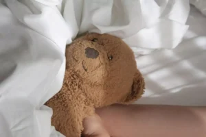 Young child sleeping with a teddy bear. The image shows only a teddy bear's head and the arms of a child holding it under the covers.