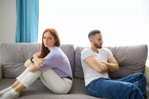 A man and a woman sit on a couch, arms folded, angrily turned away from each other. Have you ever wondered if you and your spouse are under spiritual attack or if your spouse is just a jerk?