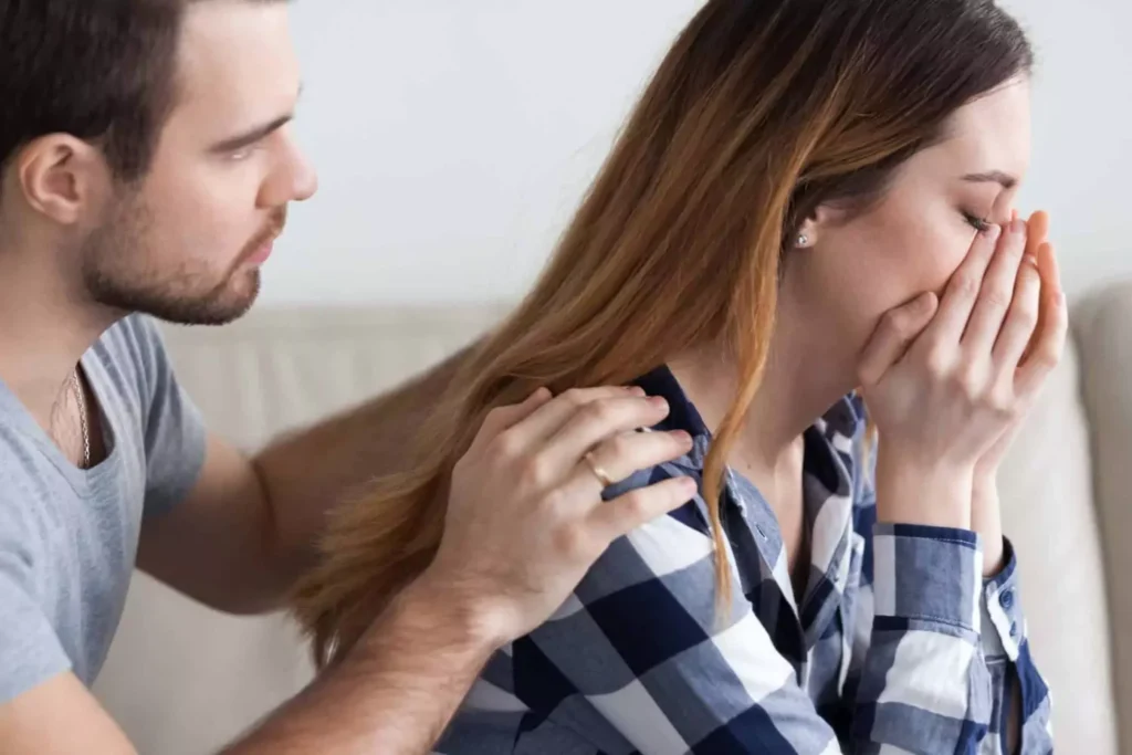 A husband comforting his grieving wife as she sits on a couch with her hands covering her face, illustrating how to support a grieving spouse with empathy and care.