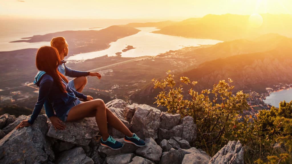 a man and woman are sitting on a mountainside and watching the sunset