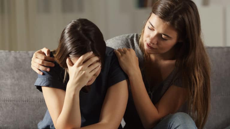 Two young women sitting on a couch. One is leaning forward crying, the other is trying to comfort her.