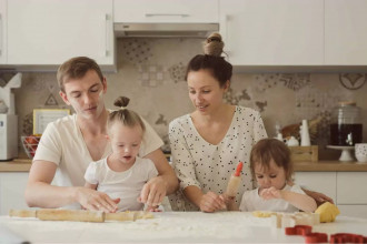 A loving family of four is gathered in their kitchen, baking together. The father, with flour on his face, helps his young daughter, who has Down syndrome, shape dough on the table. The mother smiles as she guides their other child, who is focused on rolling dough. Their teamwork and joy reflect the unique dynamics and love within a family raising special needs children.