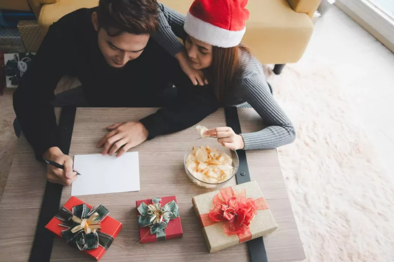 A couple sits together at a table, setting New Year’s goals together as they write on a piece of paper surrounded by festive gifts and holiday decorations.